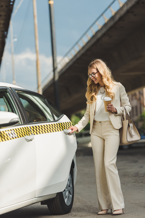 smiling blonde woman with paper cup opening door of taxi cabの写真素材