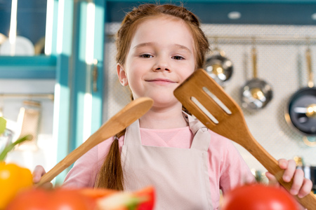 beautiful happy child in apron holding wooden utensils and smiling at cameraの写真素材