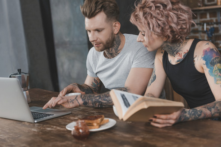 tattooed couple using laptop and reading book during breakfast in kitchenの写真素材