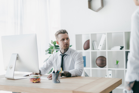handsome young tattooed businessman looking away while working with desktop computer in officeの写真素材