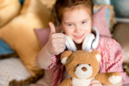 cute little child in headphones holding teddy bear and showing thumb upの写真素材