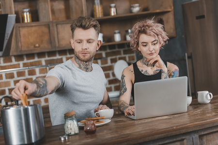young tattooed couple using laptop during breakfast in kitchenの写真素材