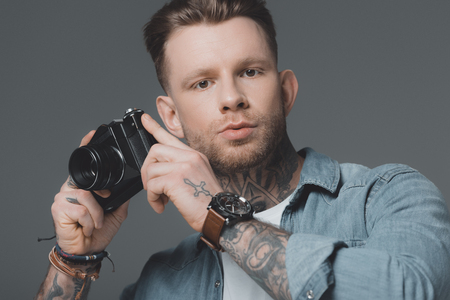 handsome young man with tattoos holding camera and looking at camera isolated on greyの写真素材