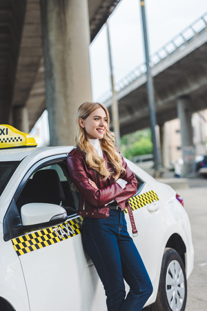 beautiful blonde girl standing with crossed arms near taxi cab and looking awayの写真素材