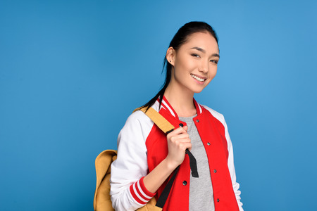 portrait of smiling asian student with backpack isolated on blue