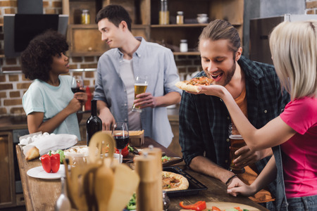 girl feeding friend with piece of pizza in kitchenの写真素材
