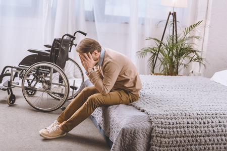 upset man with disability sitting on bed and covering face with handsの写真素材