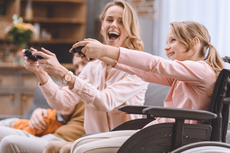 cheerful family with disabled child in wheelchair playing with joysticks together at homeの写真素材