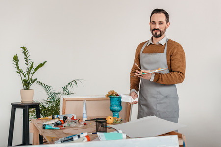 handsome mature male artist in apron holding palette and smiling at camera