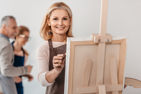beautiful mature woman smiling at camera while painting on easel at art classの写真素材