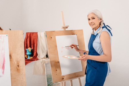 beautiful senior woman smiling at camera while painting on easel in art studioの写真素材