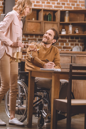 cropped shot of smiling woman holding wine glasses and bottle while husband in wheelchair taking notes at homeの写真素材