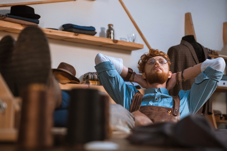 selective focus of young male fashion designer in apron and eyeglasses sitting with hands behind head at workplaceの写真素材
