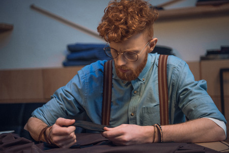 young male fashion designer in eyeglasses working with fabric at table