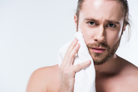 Young bearded  man looking to camera while holding bath towel in hand close to his cheek,  isolated on whiteの写真素材