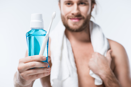 Smiling man with towel around his neck holding  tooth rinse and toothbrush in hand, isolated on whiteの写真素材