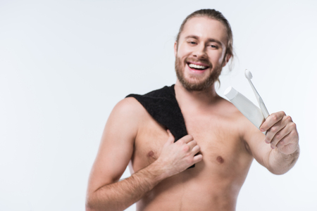 Smiling young man with black towel on shoulder holding toothpaste and toothbrush in hand, isolated on whiteの写真素材