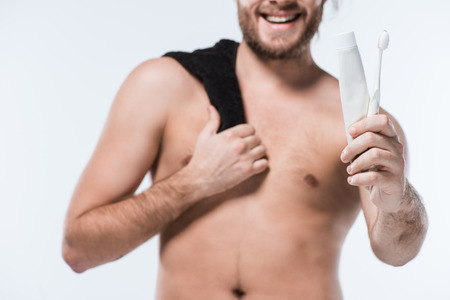 Cropped image of caucasian bearded man with black towel on shoulder holding toothpaste and toothbrush, isolated on whiteの写真素材