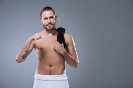 man with black towel on shoulder holding toothbrush with toothpaste in hand against his face, isolated on grayの写真素材