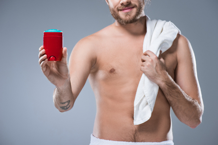 Smiling half naked man holding bath towel over shoulder in one hand and deodorant stick in another, isolated on grayの写真素材