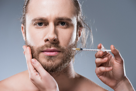 Caucasian brunette man looking at camera while persons hands making injection in his cheek, isolated on grayの写真素材