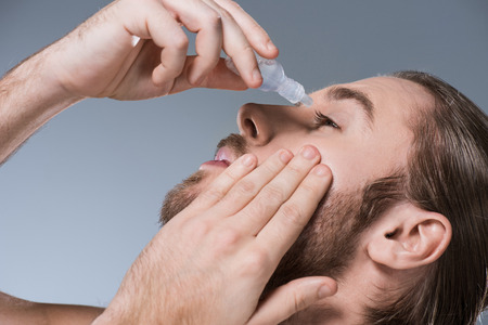 Portrait of handsome young man dripping eye drops with hand on cheek, isolated on grayの写真素材