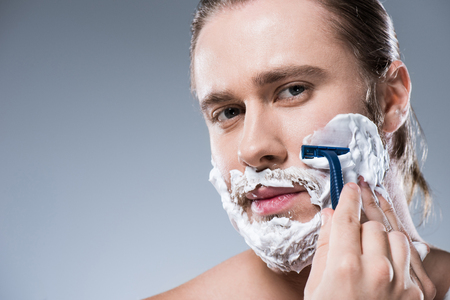 Caucasian man shaving his face with foam on his face and razor in hand, isolated on grayの写真素材
