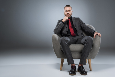 Serious young businessman sitting in armchair with hand to chin and looking to camera, isolated on grayの写真素材
