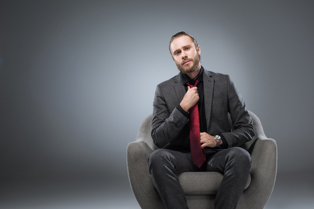 Serious businessman sitting in armchair and straightening tie, while looking at camera, isolated on grayの写真素材