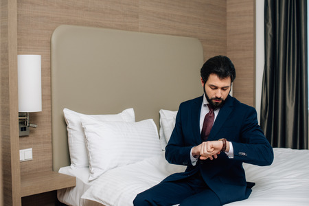 handsome businessman sitting on bed at hotel room and looking at watchの写真素材