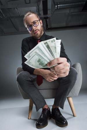 Serious businessman in glasses sitting in armchair with money in hands and looking at cameraの写真素材