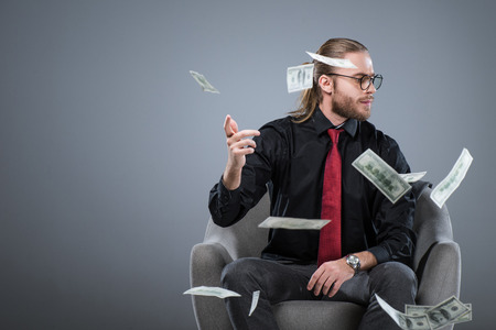 Businessman sitting in armchair while throwing money away, isolated on grayの写真素材