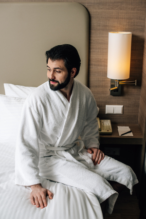 smiling man in bathrobe sitting on bed at hotel suiteの写真素材