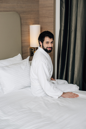 happy man in bathrobe sitting on bed at hotel suiteの写真素材