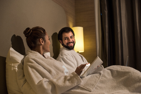 happy couple in bathrobes using devices in bed of hotel suiteの写真素材