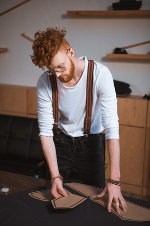 young male fashion designer in eyeglasses working with sewing patterns and fabricの写真素材