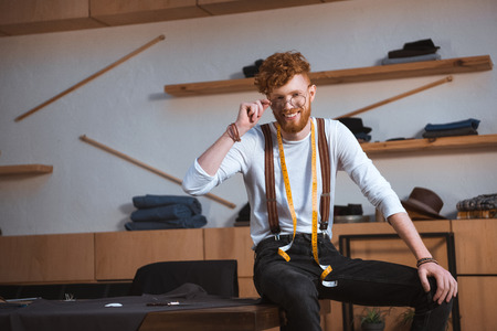 young male fashion designer in eyeglasses with measuring tape around neck smiling at camera at workplaceの写真素材