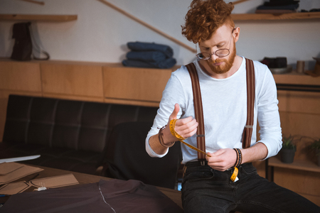 young male fashion designer in eyeglasses holding measuring tape at workshopの写真素材