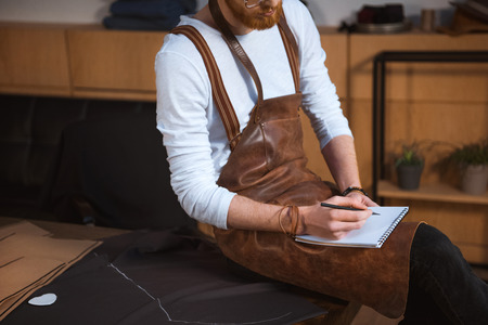cropped shot of bearded male fashion designer in apron taking notes in notebook