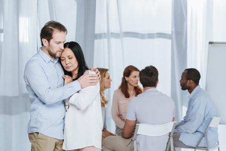 upset mature couple hugging while people sitting on chairs behind during group therapyの写真素材