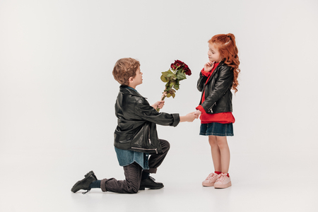 boy presenting roses bouquet to his little girlfriend while standing on knee isolated on greyの写真素材