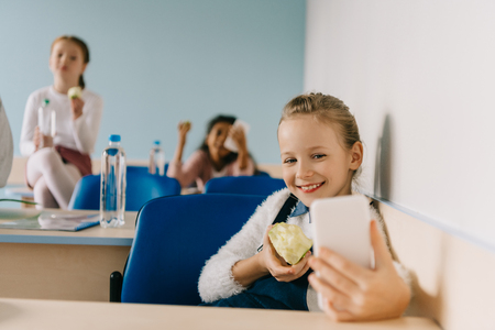 happy teen schoolgirl taking selfie at classroom while eating appleの写真素材