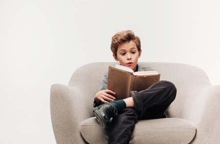 excited little schoolboy reading book in armchair isolated on greyの写真素材