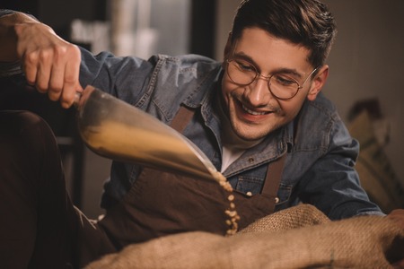 portrait of smiling worker in apron pouring coffee beans into sack bagの写真素材