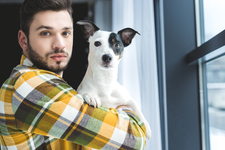 bearded man holding jack russell terrier dog and standing at windowの写真素材