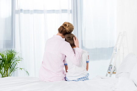 rear view of mother and son embracing while sitting on bedの写真素材