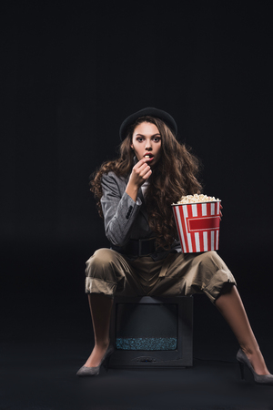 shocked young woman eating popcorn while sitting on tv and looking at camera on blackの写真素材