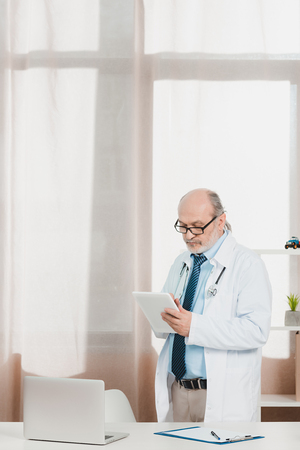 portrait of senior doctor in white coat with tablet in hands at workplace in clinicの写真素材