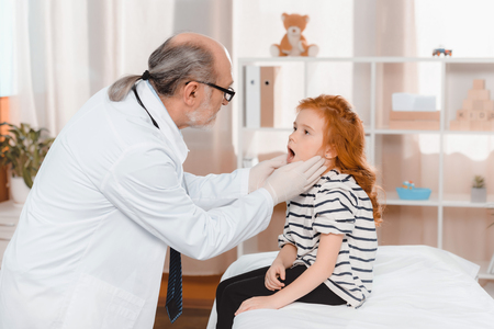 senior doctor in medical gloves examining little patients throat in clinicの写真素材