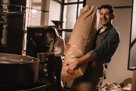 smiling man holding paper bag with coffee beans with colleague near by in coffee shopの写真素材